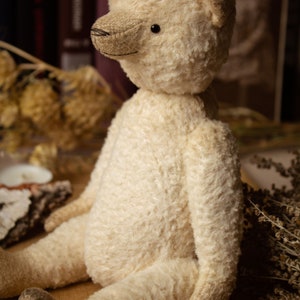 May include: A cream-colored teddy bear with black eyes sits on a brown surface with wooden beads. The bear has a stitched nose and a fluffy texture.
