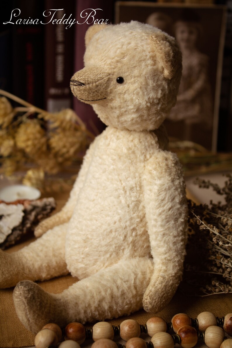 May include: A cream-colored teddy bear with black eyes sits on a brown surface with wooden beads. The bear has a stitched nose and a fluffy texture.