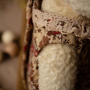 Puede incluir: Primer plano de un oso de peluche blanco que lleva un vestido floral marr&oacute;n y rojo con ribete de encaje. El vestido tiene un estampado floral marr&oacute;n y rojo.
