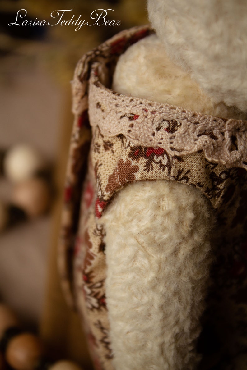 Puede incluir: Primer plano de un oso de peluche blanco que lleva un vestido floral marr&oacute;n y rojo con ribete de encaje. El vestido tiene un estampado floral marr&oacute;n y rojo.