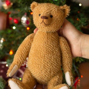 May include: A small, golden-brown teddy bear with textured fur, featuring a dark brown stitched nose and mouth. The bear has light-coloured, smooth fabric patches on the soles of its feet and the palms of its hands, with small black eyes.