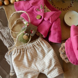 Pu&ograve; includere: Un completo per bambole fatto a mano con una giacca rosa, pantaloncini a quadri marroni e bianchi e un cappello rosa. L'abito &egrave; disposto su uno sfondo di tessuto marrone.