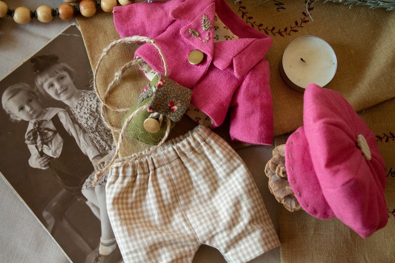 Pu&ograve; includere: Un completo per bambole fatto a mano con una giacca rosa, pantaloncini a quadri marroni e bianchi e un cappello rosa. L'abito &egrave; disposto su uno sfondo di tessuto marrone.
