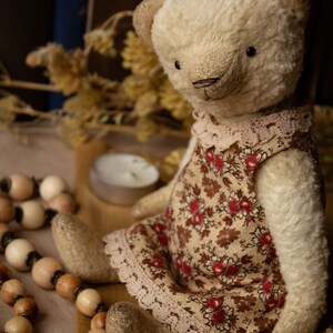 Puede incluir: Un oso de peluche de color crema que lleva un vestido floral marr&oacute;n y rojo con ribete de encaje. El oso est&aacute; sentado sobre una superficie marr&oacute;n con cuentas de madera.