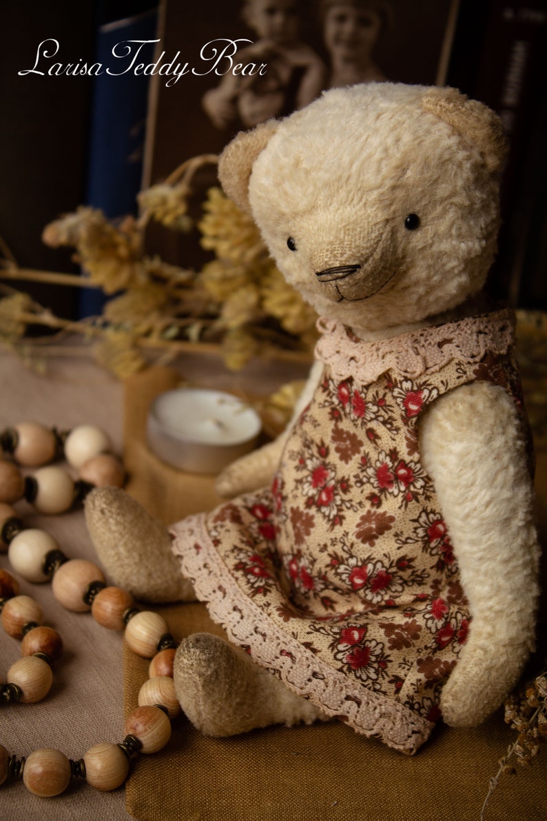 Puede incluir: Un oso de peluche de color crema que lleva un vestido floral marr&oacute;n y rojo con ribete de encaje. El oso est&aacute; sentado sobre una superficie marr&oacute;n con cuentas de madera.
