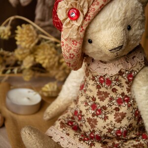 Puede incluir: Un oso de peluche blanco que lleva un vestido de estampado floral rojo y blanco y un sombrero de estampado floral rojo y blanco con una flor beige y un bot&oacute;n de strass. El oso est&aacute; sentado sobre una superficie marr&oacute;n con un collar de cuentas de madera.