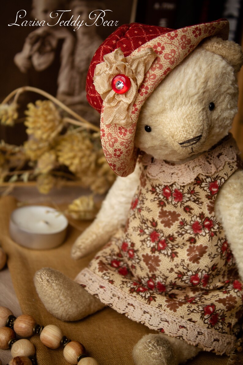 Puede incluir: Un oso de peluche blanco que lleva un vestido de estampado floral rojo y blanco y un sombrero de estampado floral rojo y blanco con una flor beige y un bot&oacute;n de strass. El oso est&aacute; sentado sobre una superficie marr&oacute;n con un collar de cuentas de madera.