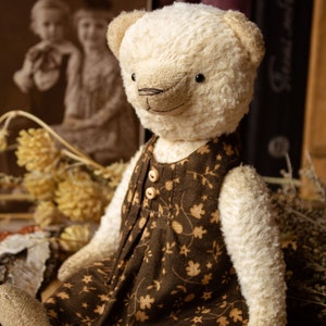 May include: A white teddy bear wearing a brown dress with a floral pattern. The bear is sitting on a brown surface with a wooden bead necklace and a candle.