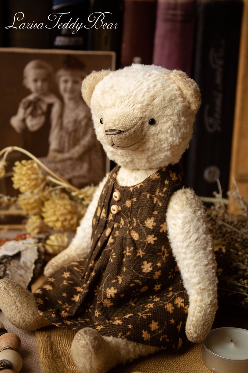 May include: A white teddy bear wearing a brown dress with a floral pattern. The bear is sitting on a brown surface with a wooden bead necklace and a candle.