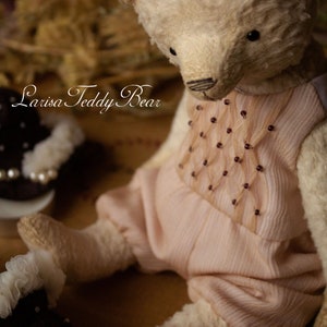 May include: A vintage white teddy bear wearing a pink dress with a beaded design. The bear is sitting on a brown surface. The text "Larisa Teddy Bear" is visible in the image.