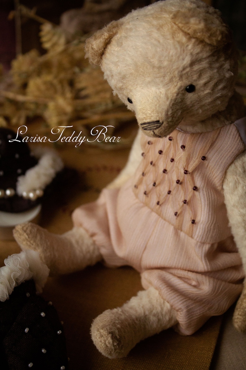 May include: A vintage white teddy bear wearing a pink dress with a beaded design. The bear is sitting on a brown surface. The text "Larisa Teddy Bear" is visible in the image.