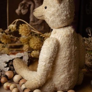 May include: A white teddy bear with a cream-colored fur, sitting on a brown surface with wooden beads. The bear has a sweet expression and is looking to the right. The bear is a vintage style teddy bear.