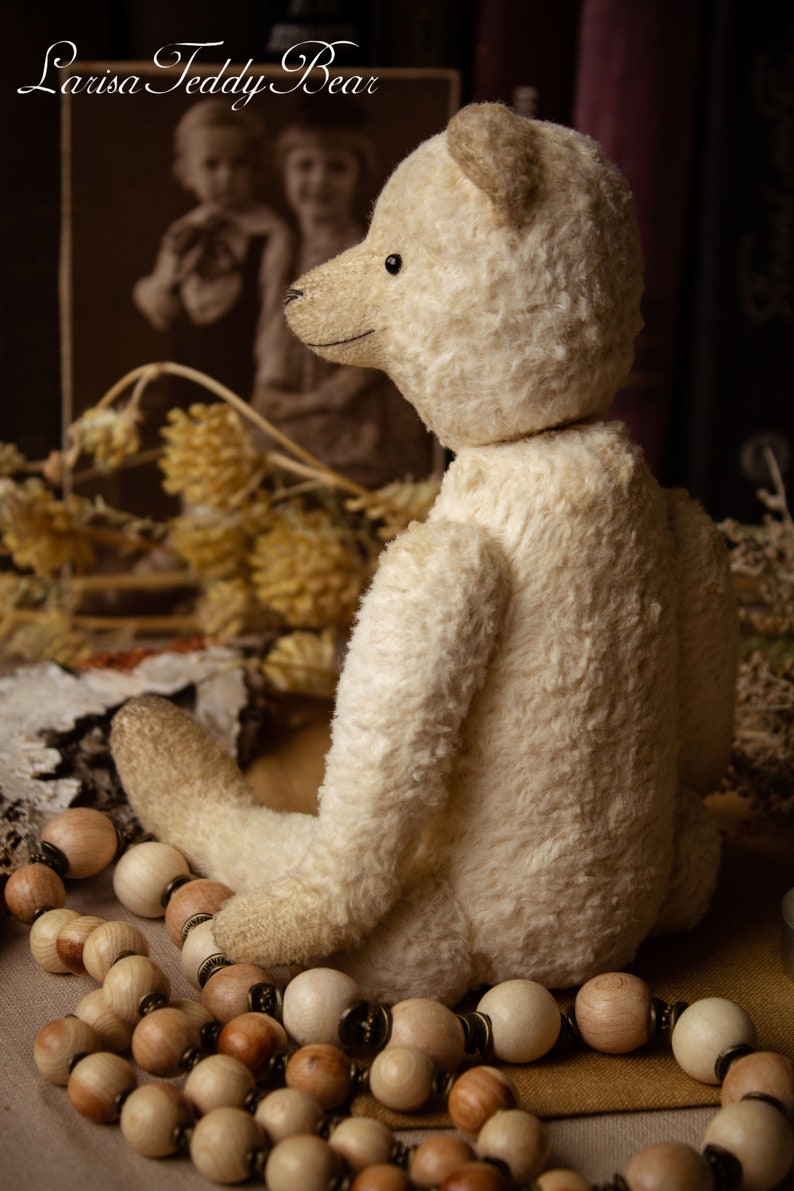 May include: A white teddy bear with a cream-colored fur, sitting on a brown surface with wooden beads. The bear has a sweet expression and is looking to the right. The bear is a vintage style teddy bear.