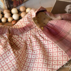 May include: A red and white plaid fabric doll dress with a brown collar and gold buttons. The dress has a diamond pattern on the front and a plaid pattern on the back.