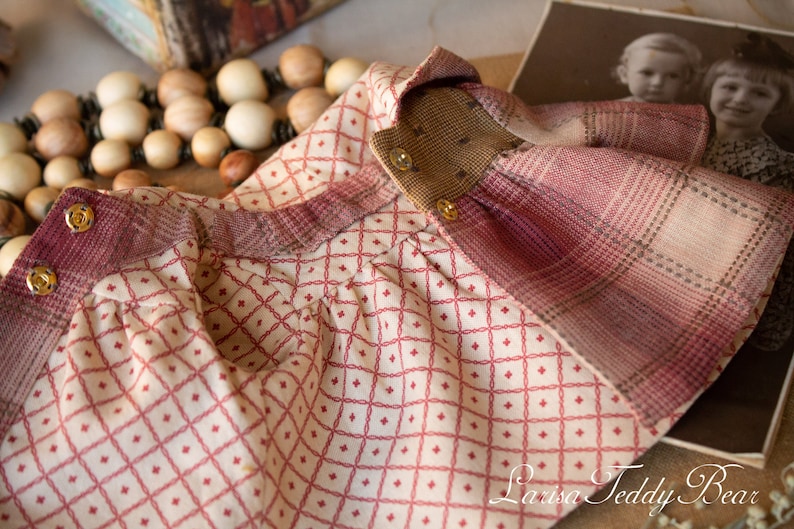 May include: A red and white plaid fabric doll dress with a brown collar and gold buttons. The dress has a diamond pattern on the front and a plaid pattern on the back.