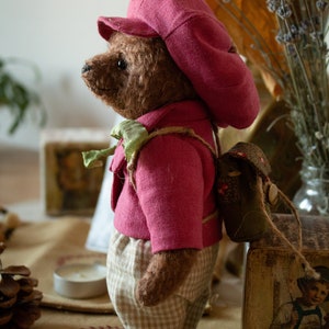 Pu&ograve; includere: Un orsacchiotto di peluche marrone che indossa un cappello rosa e una giacca rosa con un papillon verde. L'orso indossa pantaloni a quadri marroni e bianchi e un piccolo zaino verde. L'orso &egrave; in piedi su una superficie marrone.