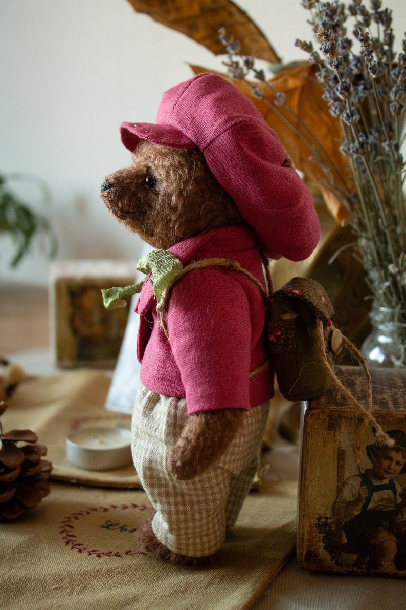 Pu&ograve; includere: Un orsacchiotto di peluche marrone che indossa un cappello rosa e una giacca rosa con un papillon verde. L'orso indossa pantaloni a quadri marroni e bianchi e un piccolo zaino verde. L'orso &egrave; in piedi su una superficie marrone.