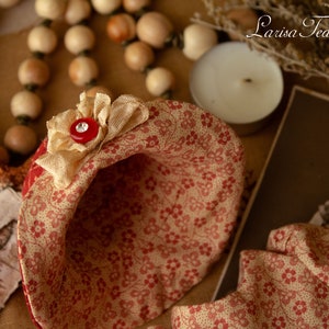 Puede incluir: Un sombrero de tela rojo y crema con un patr&oacute;n floral y un bot&oacute;n rojo. El sombrero est&aacute; sobre una superficie de madera con un collar de cuentas y una vela.