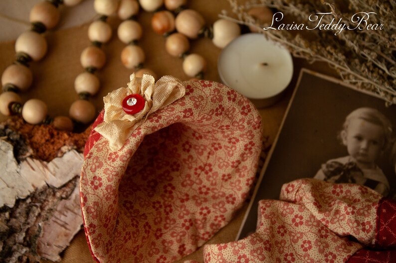 Puede incluir: Un sombrero de tela rojo y crema con un patr&oacute;n floral y un bot&oacute;n rojo. El sombrero est&aacute; sobre una superficie de madera con un collar de cuentas y una vela.
