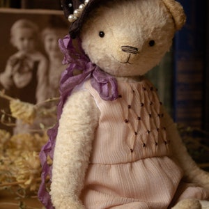 May include: A handmade teddy bear wearing a brown hat with pearl accents and a pink dress with purple ribbon accents. The bear is sitting on a wooden surface with a candle in the foreground.