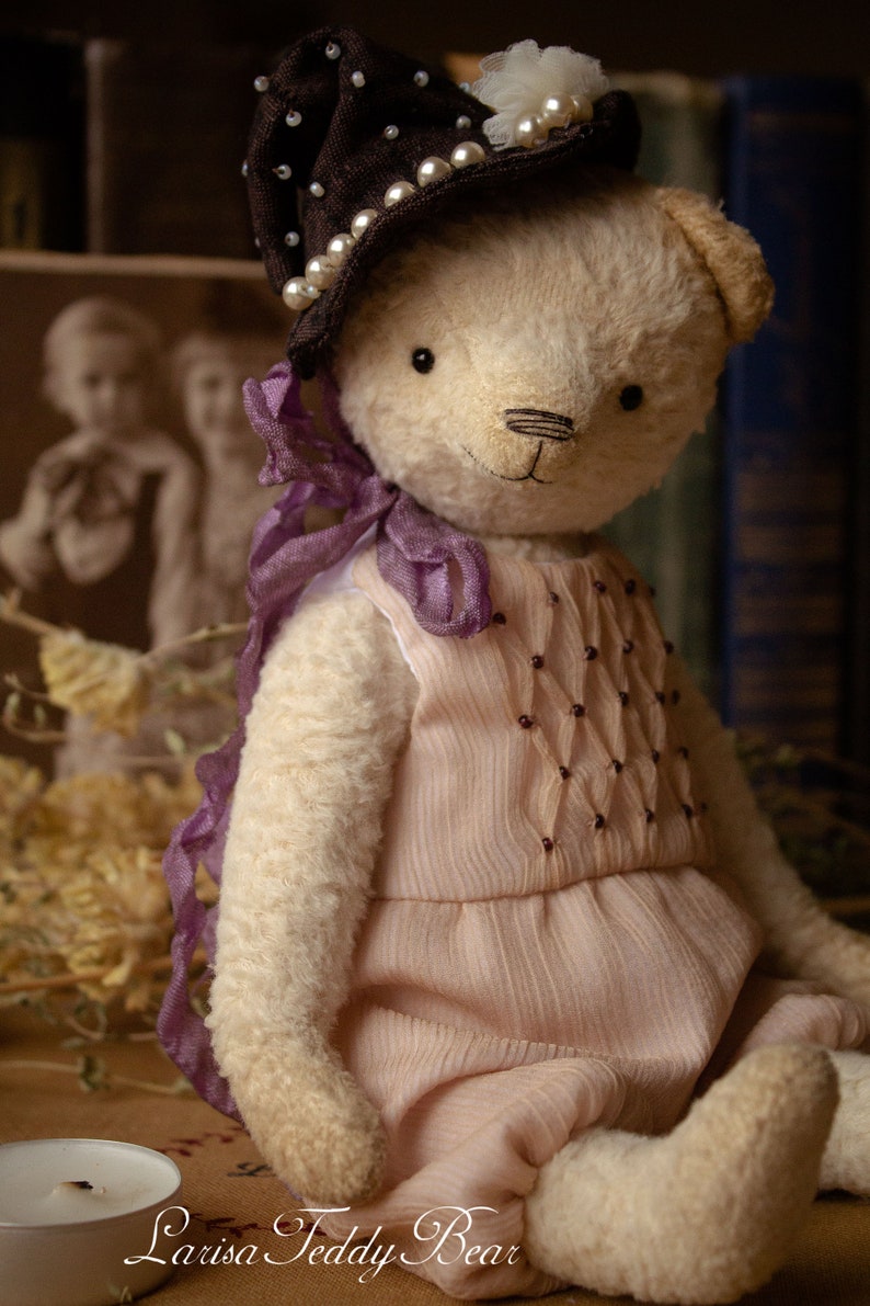 May include: A handmade teddy bear wearing a brown hat with pearl accents and a pink dress with purple ribbon accents. The bear is sitting on a wooden surface with a candle in the foreground.