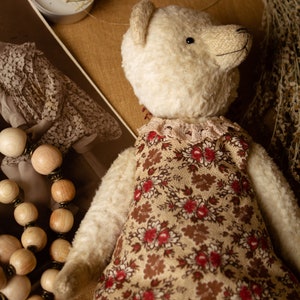 Puede incluir: Un oso de peluche blanco que lleva un vestido floral marr&oacute;n y blanco con ribete de encaje. El oso est&aacute; tumbado sobre una superficie de tela marr&oacute;n con flores secas y una vela en el fondo.