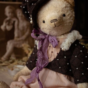 May include: A handmade teddy bear wearing a brown hat with white pearl accents, a purple ribbon, and a brown jacket with white pearl accents. The bear is sitting on a light brown surface.