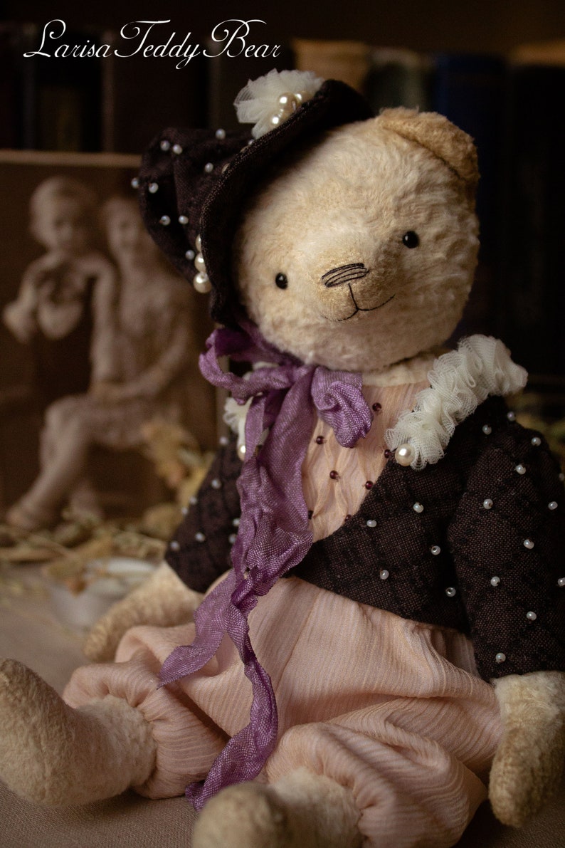 May include: A handmade teddy bear wearing a brown hat with white pearl accents, a purple ribbon, and a brown jacket with white pearl accents. The bear is sitting on a light brown surface.