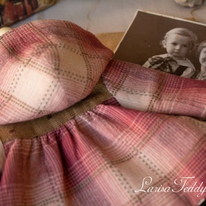 May include: A red and white plaid dress with a matching hood. The dress is made of a soft, textured fabric. The dress is laying on a table with a vintage photo of two children in the background. The photo has the text "Larisa Teddy Bear" at the bottom.