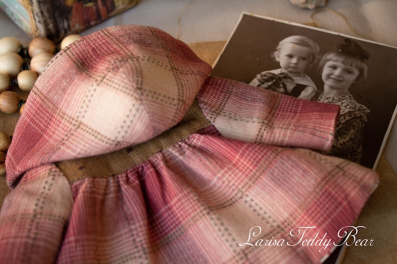 May include: A red and white plaid dress with a matching hood. The dress is made of a soft, textured fabric. The dress is laying on a table with a vintage photo of two children in the background. The photo has the text "Larisa Teddy Bear" at the bottom.