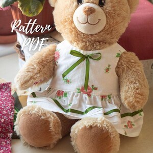 May include: A tan plush teddy bear wearing a white sleeveless dress with a small pink rose floral pattern, a green ribbon bow at the neckline, and a green ribbon trim along the hem. The image includes the white text Pattern PDF.