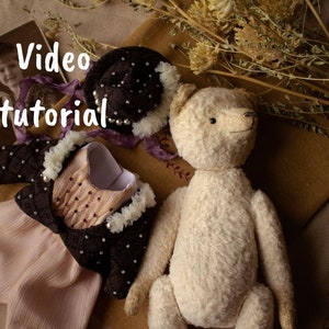 May include: A white teddy bear with a brown and white dress and a black hat with white beads. The text "Video tutorial" is written in white on a black background.
