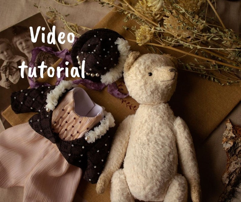 May include: A white teddy bear with a brown and white dress and a black hat with white beads. The text "Video tutorial" is written in white on a black background.