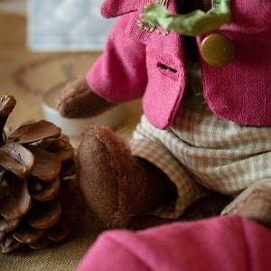 Pu&ograve; includere: Un orsacchiotto di peluche marrone che indossa una giacca rosa con un papillon verde e pantaloni marroni con un motivo a quadri bianchi e marroni. L'orso &egrave; seduto su una superficie marrone.