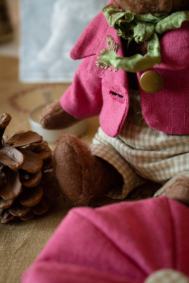 Pu&ograve; includere: Un orsacchiotto di peluche marrone che indossa una giacca rosa con un papillon verde e pantaloni marroni con un motivo a quadri bianchi e marroni. L'orso &egrave; seduto su una superficie marrone.