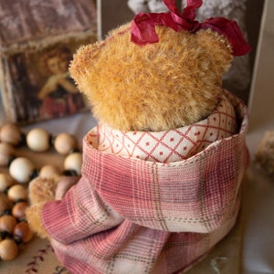 May include: A plush teddy bear wearing a red and white plaid jacket with a red bow on its head. The bear is sitting on a brown surface with a wooden bead necklace and a candle in the foreground.