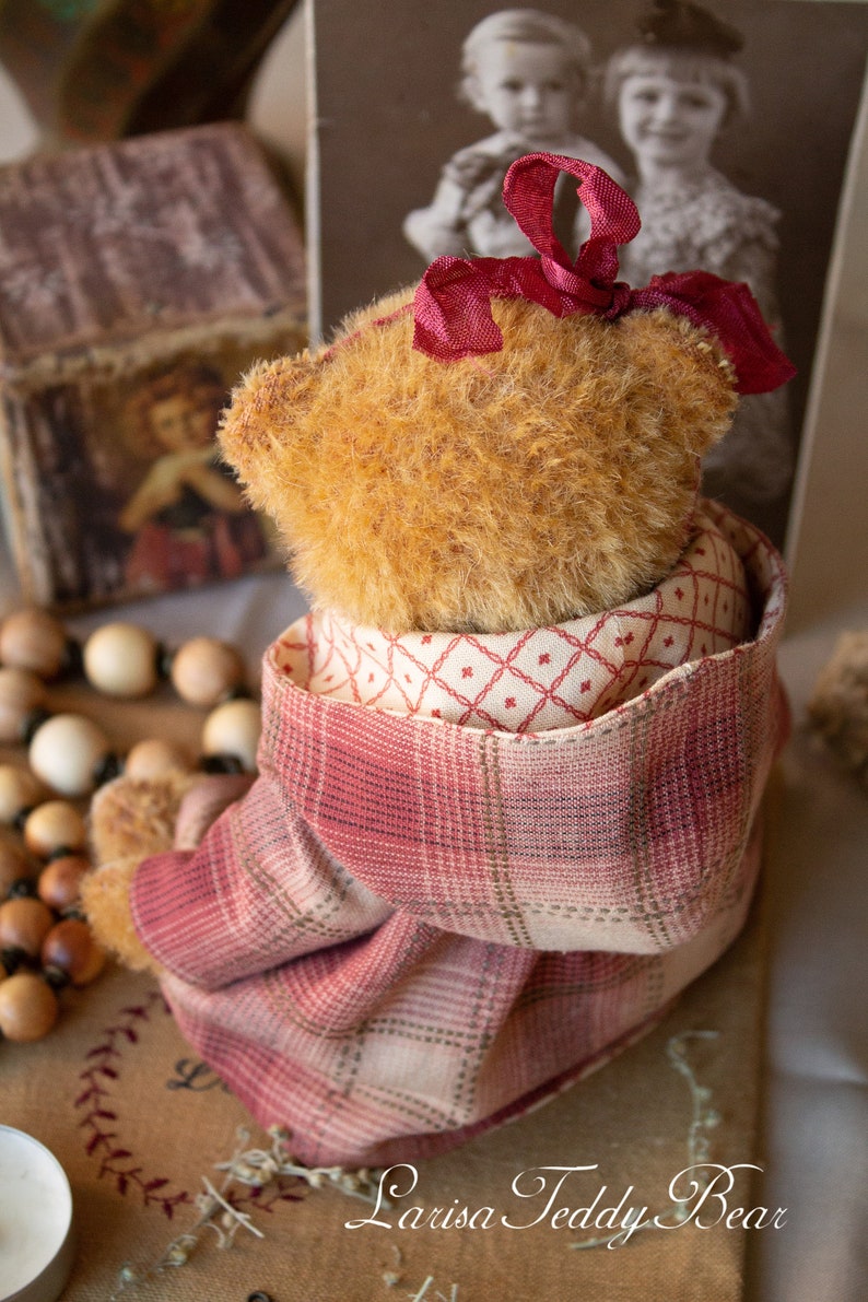 May include: A plush teddy bear wearing a red and white plaid jacket with a red bow on its head. The bear is sitting on a brown surface with a wooden bead necklace and a candle in the foreground.