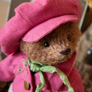 Pu&ograve; includere: Un orsacchiotto di peluche marrone che indossa un cappello rosa e una giacca rosa con una sciarpa verde. L'orso ha un naso cucito e occhi neri.