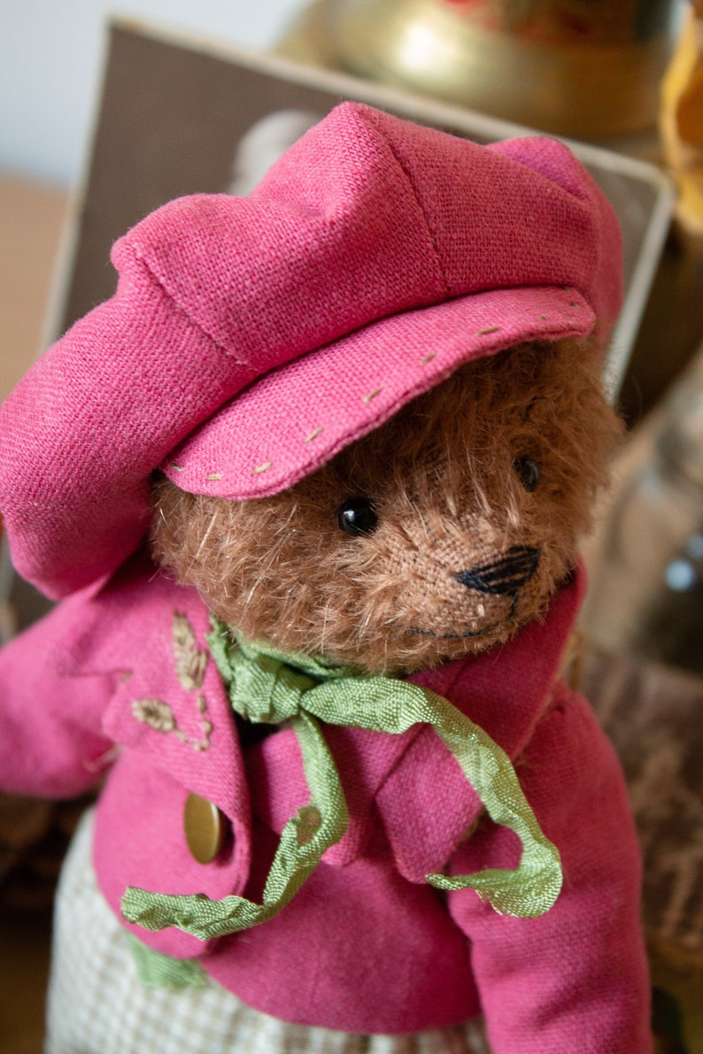 Pu&ograve; includere: Un orsacchiotto di peluche marrone che indossa un cappello rosa e una giacca rosa con una sciarpa verde. L'orso ha un naso cucito e occhi neri.