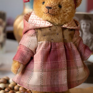 May include: A brown teddy bear wearing a red and white plaid dress with a brown hooded jacket and a red bow. The bear is standing on a brown surface with a wooden bead garland and a silver candle holder.