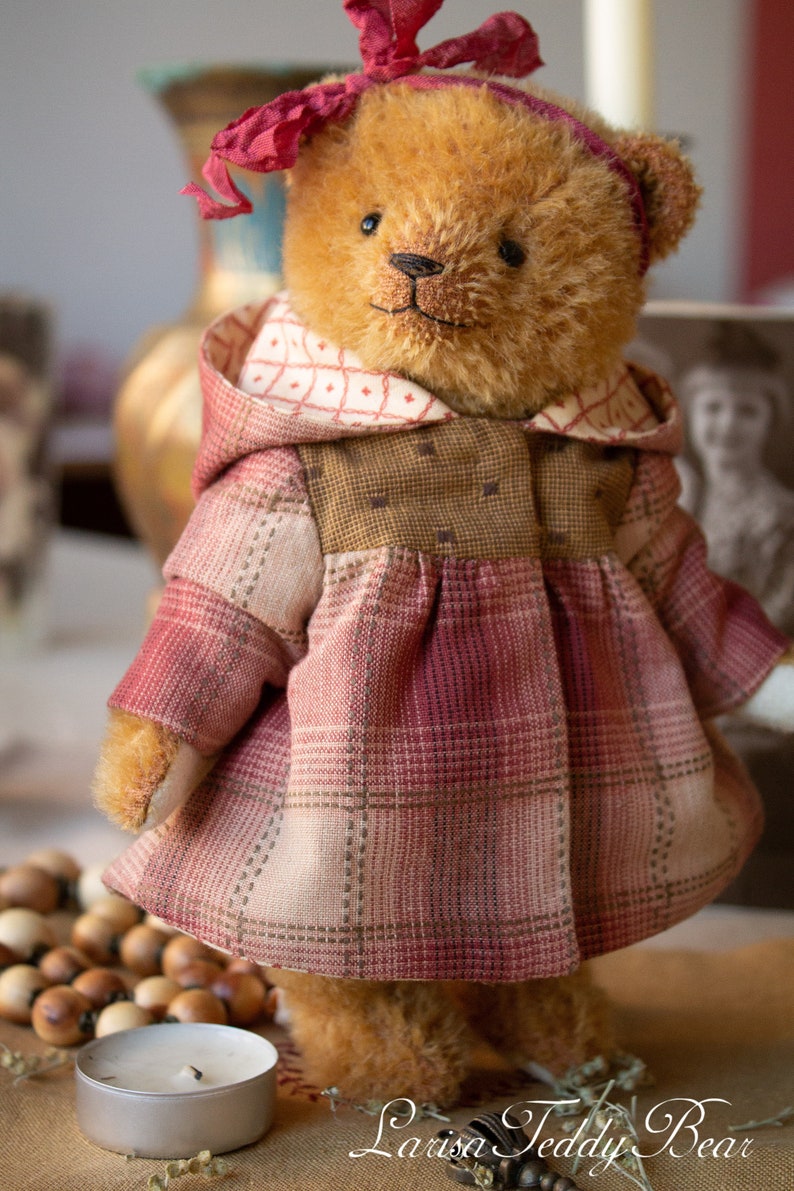 May include: A brown teddy bear wearing a red and white plaid dress with a brown hooded jacket and a red bow. The bear is standing on a brown surface with a wooden bead garland and a silver candle holder.