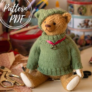 May include: A golden-brown teddy bear wearing a sage green knitted jumper and a matching hat. The bear has a dark brown nose, black eyes, and a pink ribbon around its neck with a small metal rocking horse charm. The text Pattern PDF is visible.
