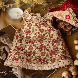 Puede incluir: Un vestido floral beige y marr&oacute;n con ribete de encaje, un sombrero rojo y blanco con una cinta marr&oacute;n y un collar de cuentas de madera. El vestido est&aacute; sobre una superficie marr&oacute;n con una foto de dos ni&ntilde;os en el fondo.
