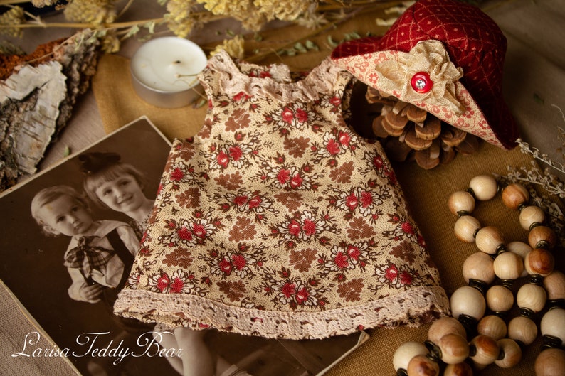 Puede incluir: Un vestido floral beige y marr&oacute;n con ribete de encaje, un sombrero rojo y blanco con una cinta marr&oacute;n y un collar de cuentas de madera. El vestido est&aacute; sobre una superficie marr&oacute;n con una foto de dos ni&ntilde;os en el fondo.