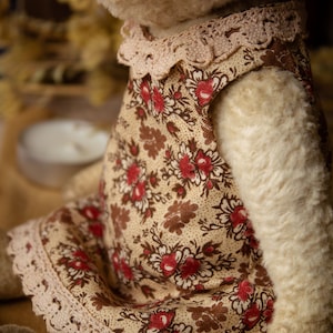 Puede incluir: Un oso de peluche blanco que lleva un vestido floral marr&oacute;n y crema con ribete de encaje. El vestido tiene un estampado floral rojo. El oso de peluche est&aacute; sentado sobre una superficie marr&oacute;n.