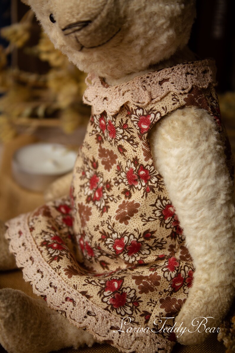 Puede incluir: Un oso de peluche blanco que lleva un vestido floral marr&oacute;n y crema con ribete de encaje. El vestido tiene un estampado floral rojo. El oso de peluche est&aacute; sentado sobre una superficie marr&oacute;n.