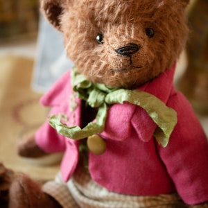 Pu&ograve; includere: Un orsacchiotto marrone che indossa una giacca rosa con una sciarpa verde e pantaloni a quadri marroni e bianchi.