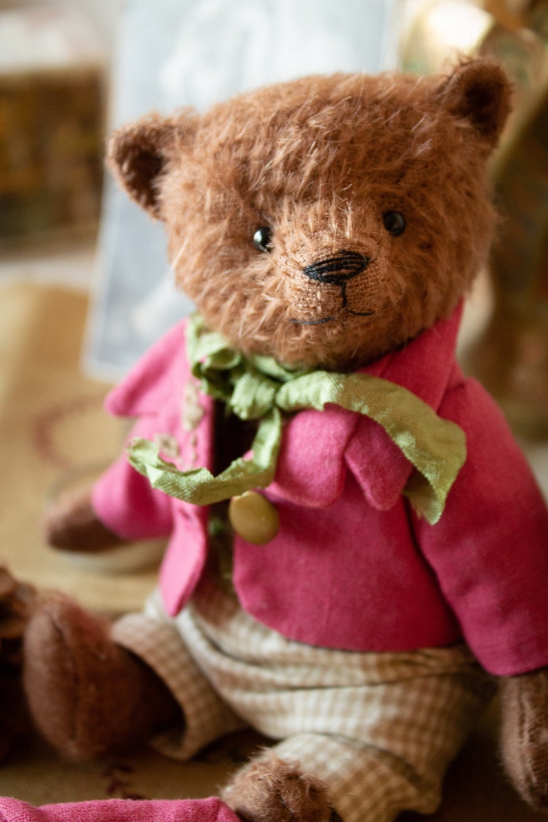 Pu&ograve; includere: Un orsacchiotto marrone che indossa una giacca rosa con una sciarpa verde e pantaloni a quadri marroni e bianchi.