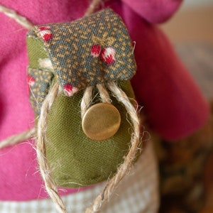 Pu&ograve; includere: Una piccola borsa di tessuto verde con una patta a motivo marrone e bianco, legata con uno spago e un bottone in ottone. La borsa &egrave; attaccata a un capo di abbigliamento in tessuto rosa.