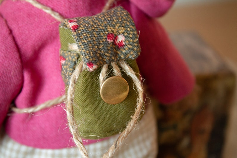 Pu&ograve; includere: Una piccola borsa di tessuto verde con una patta a motivo marrone e bianco, legata con uno spago e un bottone in ottone. La borsa &egrave; attaccata a un capo di abbigliamento in tessuto rosa.